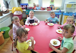 dzieci siedzą przy okrągłym stoliku i z talerzyków jedzą owocową sałatkę